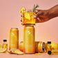 Hand holding a glass of orange drink with garnishes, surrounded by 'Vitality Ginger' products on a pink background.